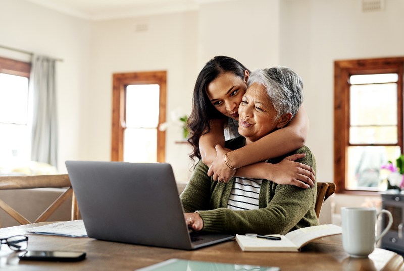 A woman hugging her mom while she is on a laptop working on her life insurance plan by the Law Offices of Robert E. O’Connor